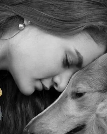 Une femme appuie son front contre son chien, dans un pull doré, en noir et blanc